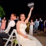 A bride and groom sitting in chairs holding up a sign.