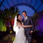 A bride and groom pose for the camera under an arch.